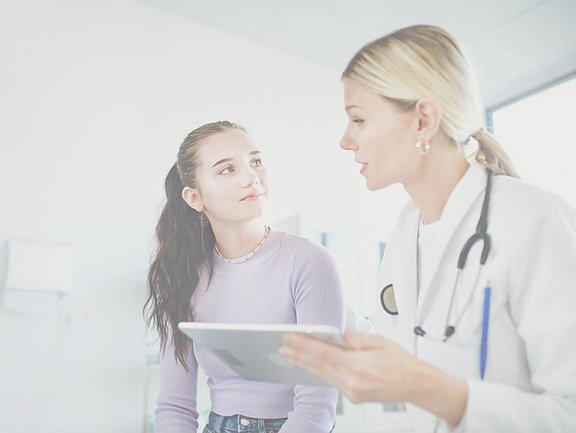 Eine Ärztin mit einem Tablet spricht mit einer jungen Frau in einer Arztpraxis. Für verständliche Erklärungen von Begriffen, die in der Allergologie verwendet werden, schaue in unser Allergie-Lexikon.